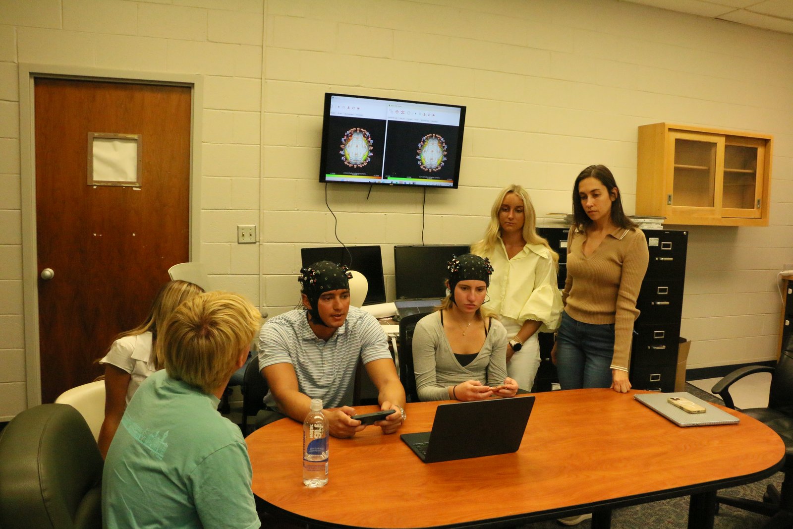 Two students wearing fNIRS caps during the PSYC 7400 hyperscanning demo, with both brains displayed on the monitor